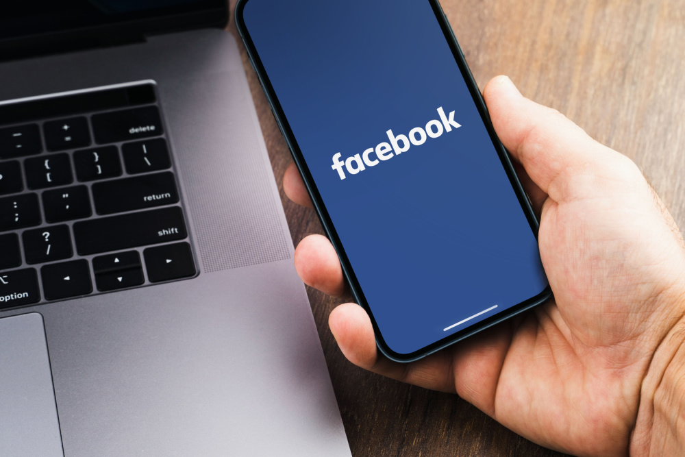 Male hand holding a smartphone with Facebook app on the screen with a computer beside it